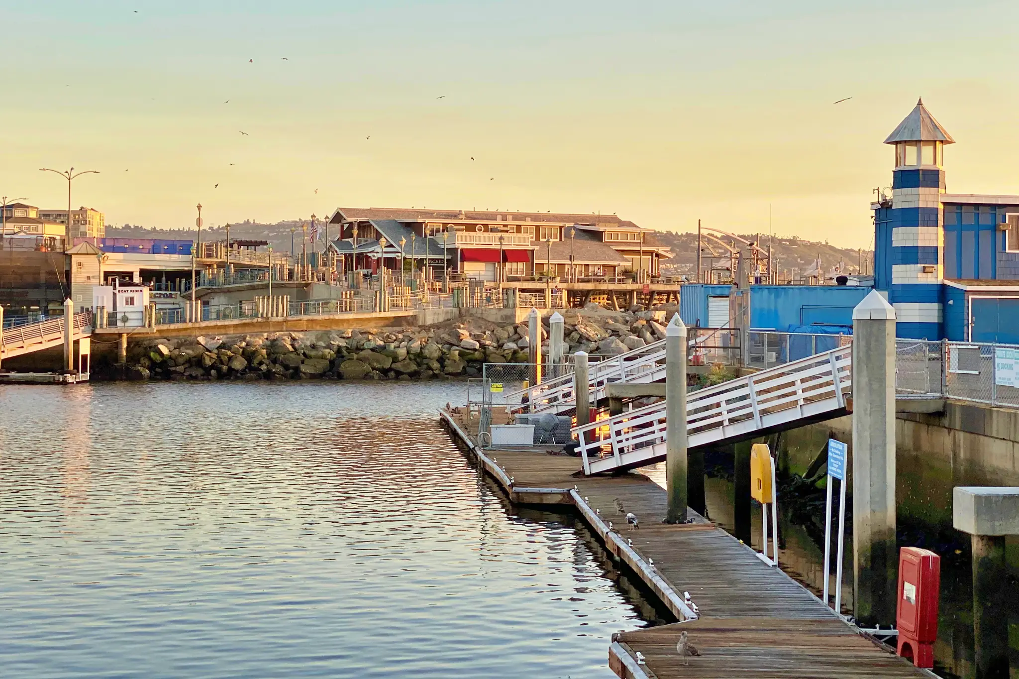 Marina dock at golden hour with wooden walkways, a small striped lighthouse, and waterfront buildings in the background, under a clear sky with birds flying