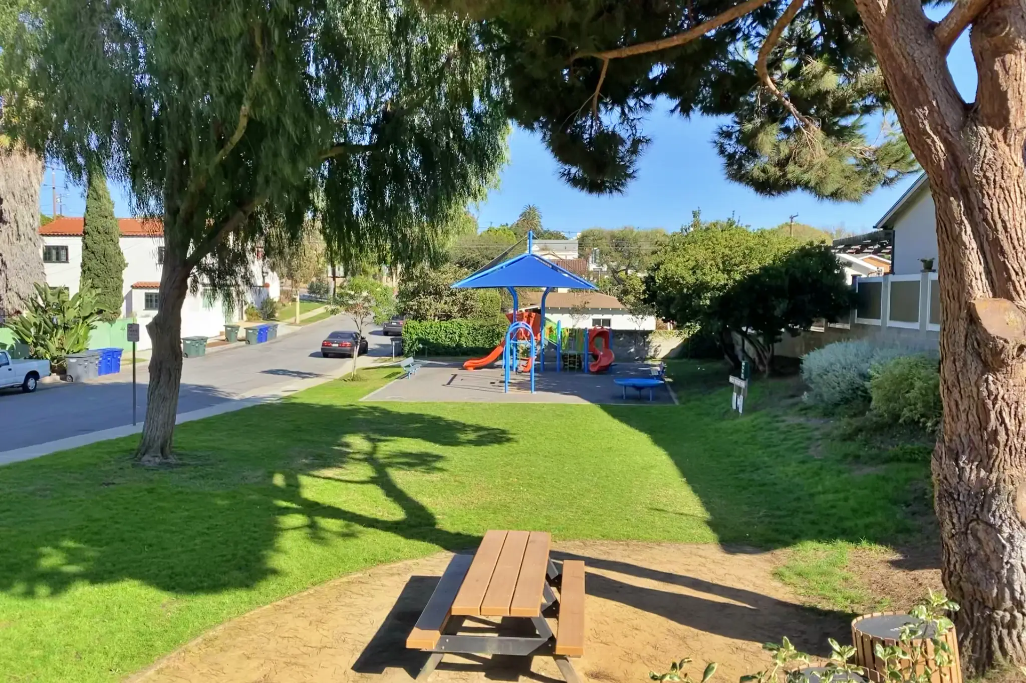 Neighborhood park in El Segundo, California with a blue-shaded playground, red and orange slides, picnic table, green grass, and surrounding residential homes