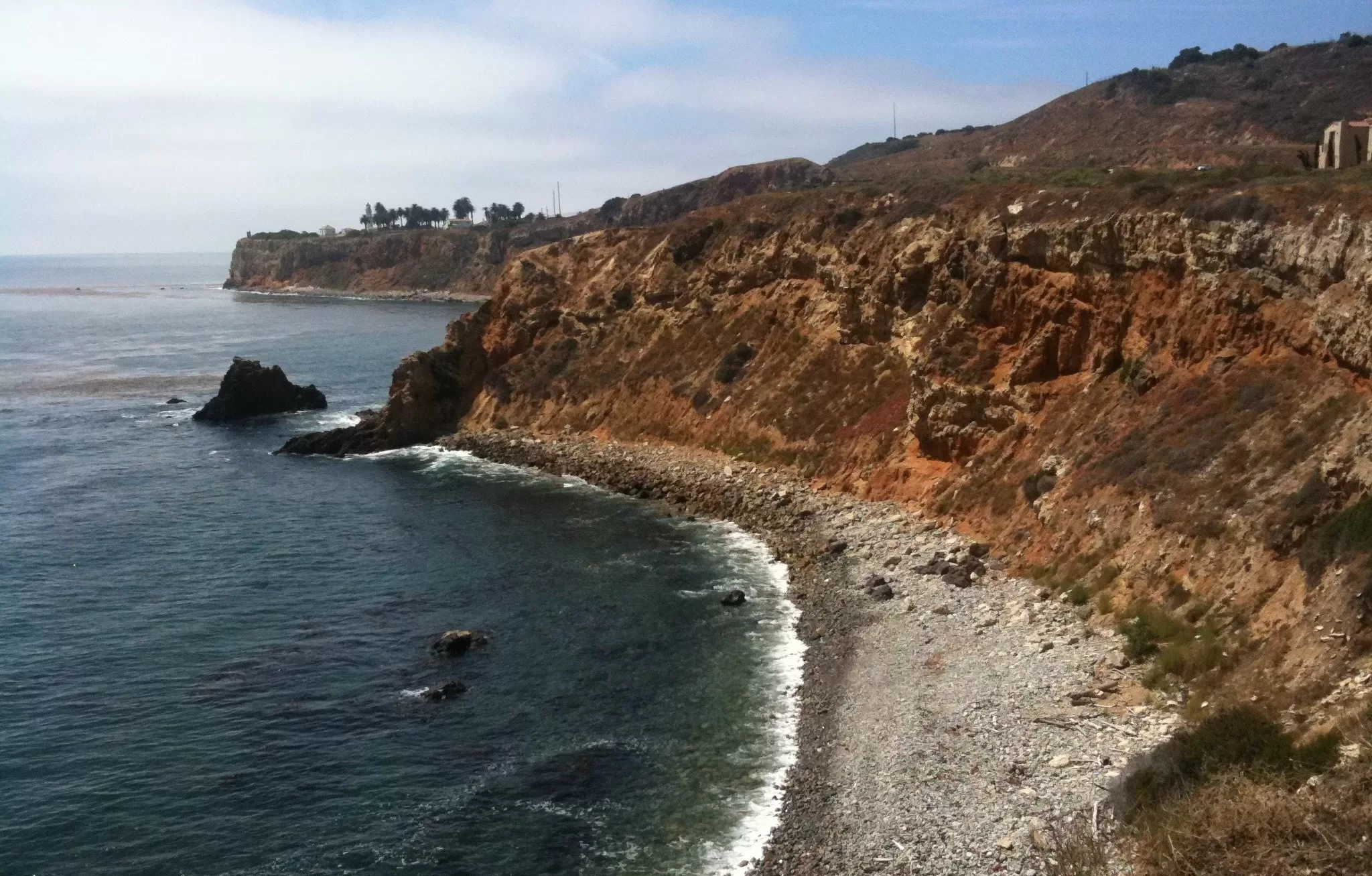 Rocky coastline with steep, rugged cliffs along a clear blue ocean under a partly cloudy sky, with sparse vegetation and a few distant buildings atop the hills